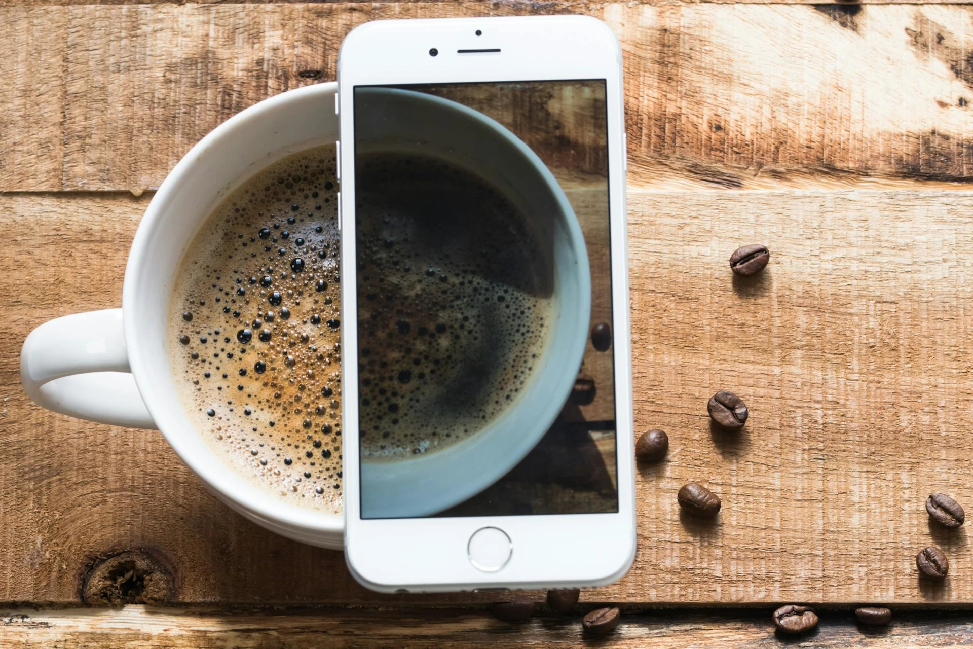 Stylish image of a cup of coffee and a smartphone on a wooden table, arranged so that the cup of partially displayed on the phone screen.