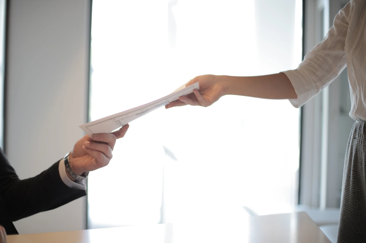 In an office, a professionally dressed woman hands a file to a professionally dressed man over a desk.
