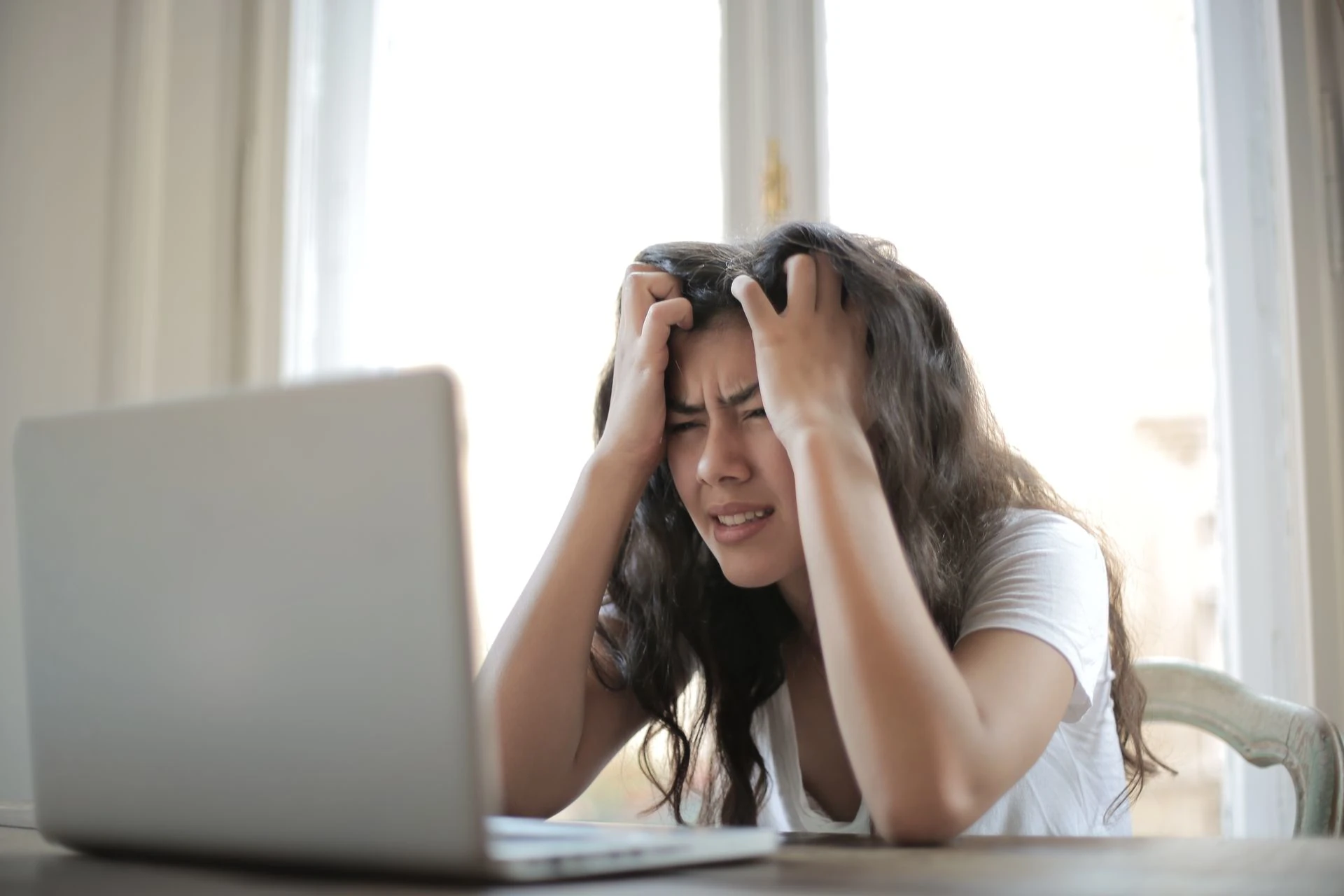 A stressed woman sits at a table holding her head in her hands while staring at a laptop.